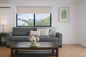 Un salón con un sofá gris, una mesa de centro de madera y una ventana con vistas a edificios y áreas verdes.