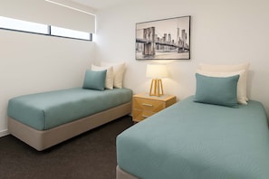 Ett hotellrum med två separata sängar, ett nattduksbord med en lampa, en inramad tavla med en stadsbild på väggen och ett fönster med persienner.
