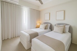 Ein Hotelzimmer mit zwei Betten, einem Deckenventilator, einem Fenster mit weißen Vorhängen und zwei gerahmten Bildern an der Wand.