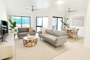 A spacious living room with a grey sofa, a round wooden coffee table, and a dining area with a table and chairs. - Townsville Waterfront Apartment with 2 BR Pool & Parking (Townsville City)