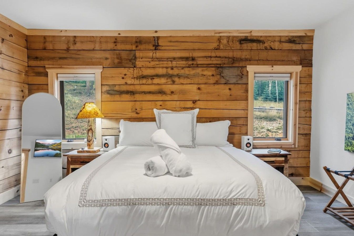 A bedroom with a wooden ceiling, a bed with white bedding, two bedside tables with lamps, and a view of trees through the window.