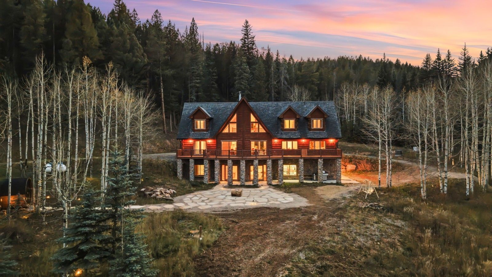 A large, multi-story log cabin with a stone foundation, surrounded by a forested landscape.