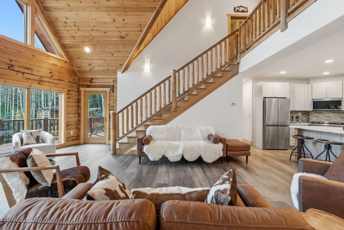 A spacious living area with a wooden staircase, a sofa with a white fur throw, and a kitchen with stainless steel appliances.