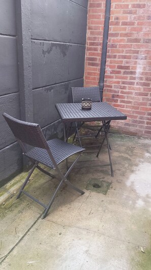 A metal table and two wicker chairs positioned against a brick wall with a metal fence. - Ready Player, jeux, wifi, proche Bollaert, 2 pers (Lens)