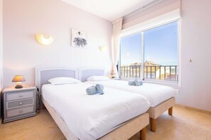 Ein Hotelzimmer mit zwei Betten, einem Nachttisch mit Lampe, einer Wandleuchte und einem großen Fenster mit Blick.