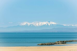 Eine Küstenlandschaft mit Sandstrand, ruhiger See und schneebedeckten Bergen im Hintergrund.