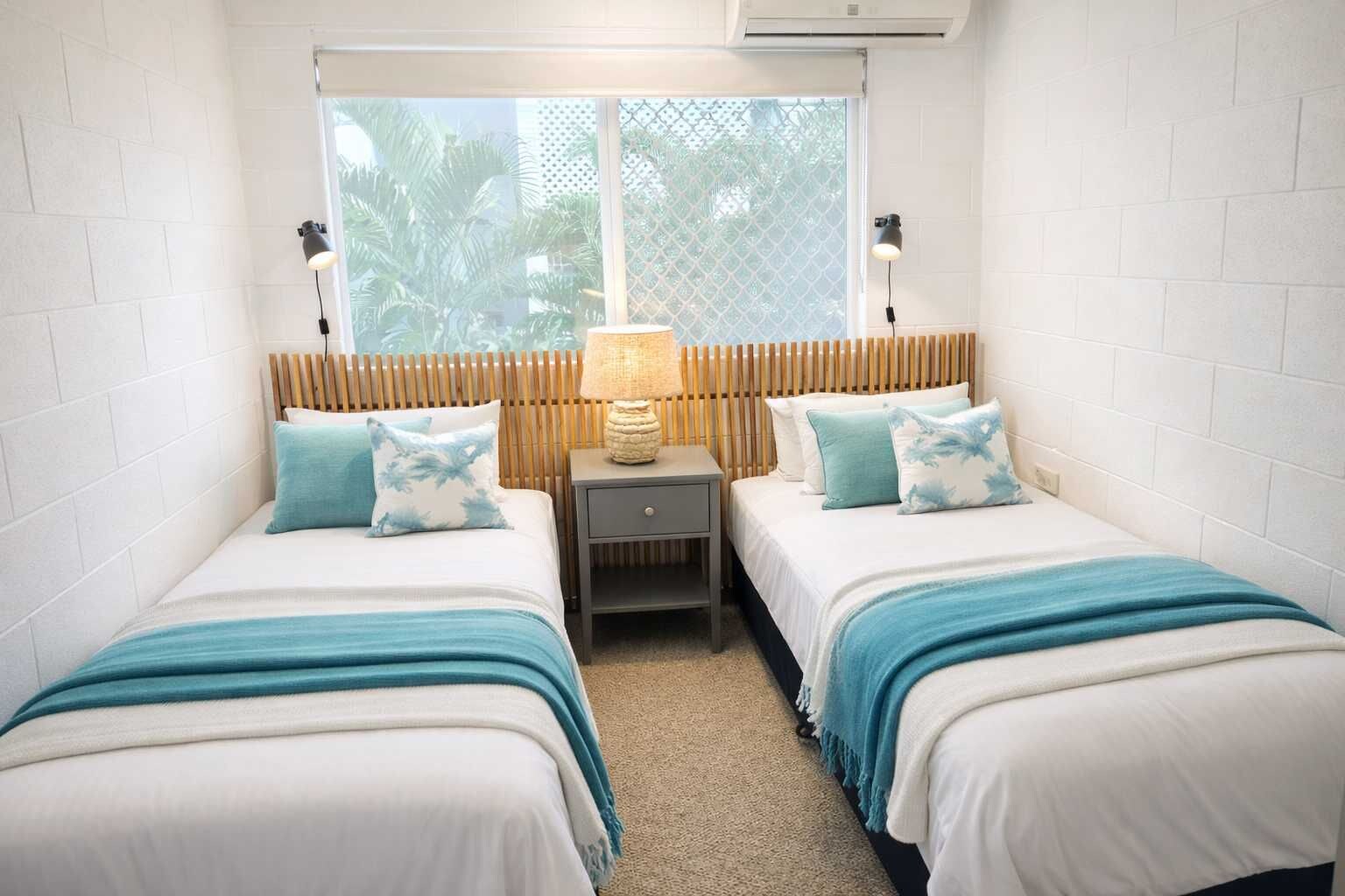 A room with two beds, a nightstand, a lamp, and a window with a view of greenery.