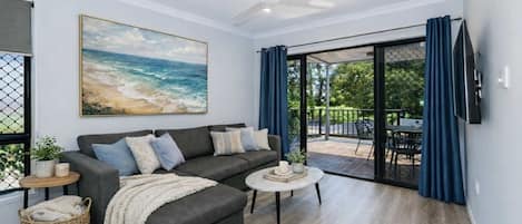 A modern living room with a grey sectional sofa, a round coffee table, a ceiling fan, and a large painting of a beach scene.
