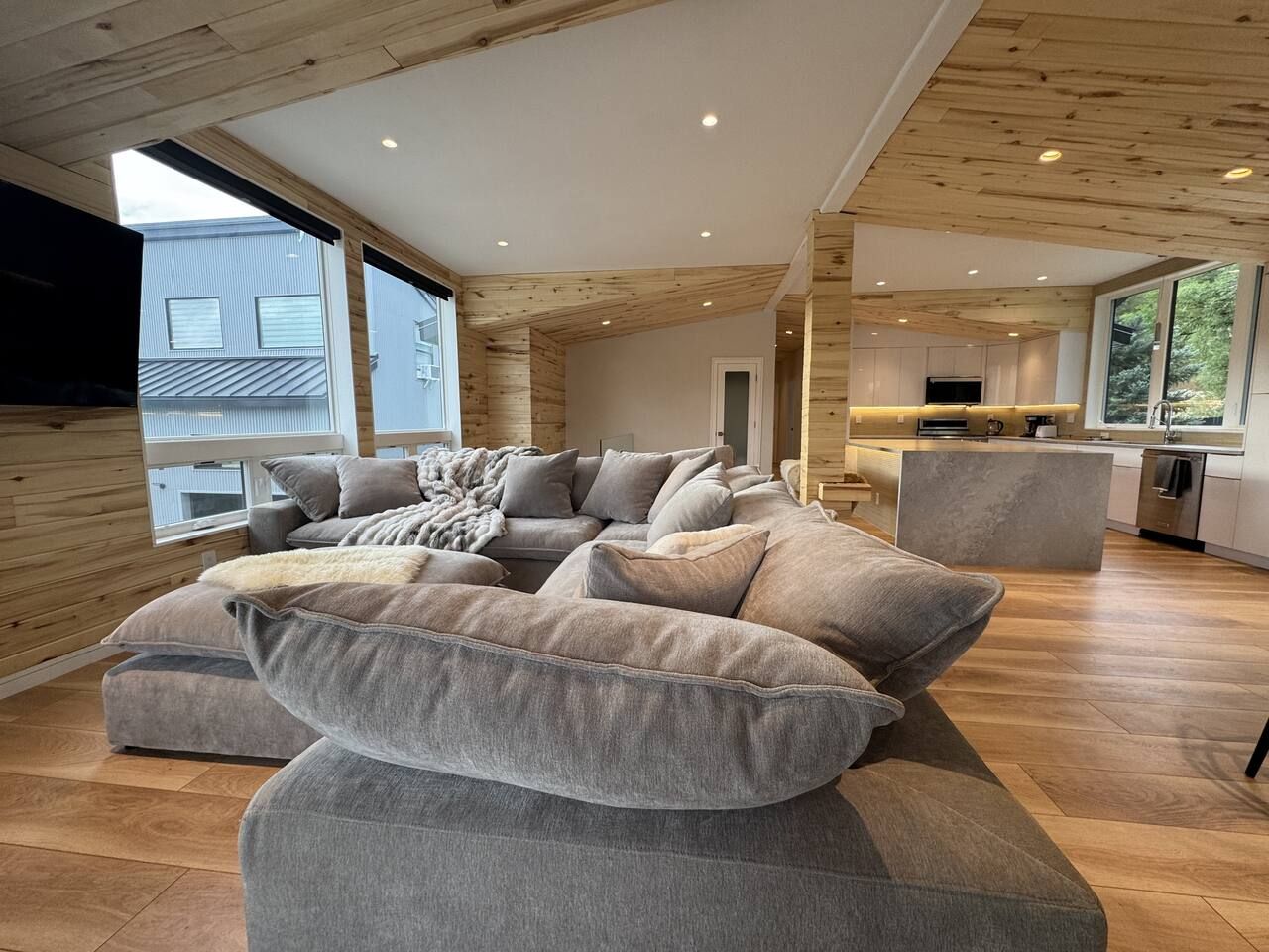 A modern living room with a large sectional sofa, wooden ceiling, and a view of a kitchen and exterior.