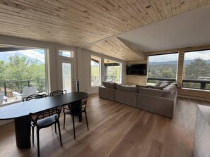 A spacious living area with a dining table, sofa, and large windows offering a view of the outdoors. - Mt. Sopris Views | Pool | 3BR Mountain Retreat (Carbondale)