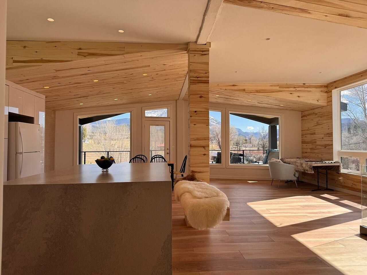 A modern interior with a wooden ceiling, a kitchen area, a dining space with chairs, and large windows offering a view of the outdoors.