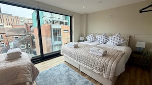 A bedroom with a large window, a bed with a quilted cover, a bedside table with a lamp, and a view of buildings outside. - Cozy Penthouse - 2 Bedroom in Central Manchester (Manchester)