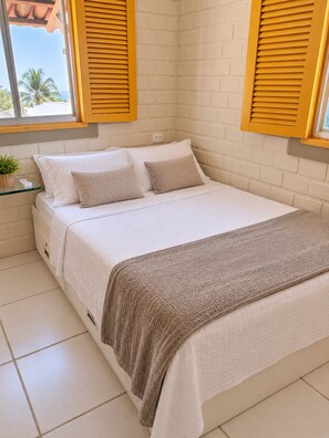 A bedroom with a bed, pillows, a bedside table with a plant, and a window with shutters. - Cozy duplex in Porto de Galinhas, 50m from the sea. • Pool • Grill (Ipojuca)