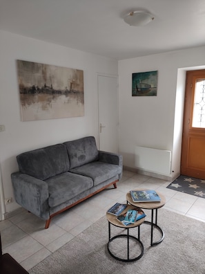 A living room with a grey sofa, a small round coffee table with books, a painting on the wall, and a door with a window. - Townhouse with a terrace and garden, 180 m from the lock. (Tinténiac)