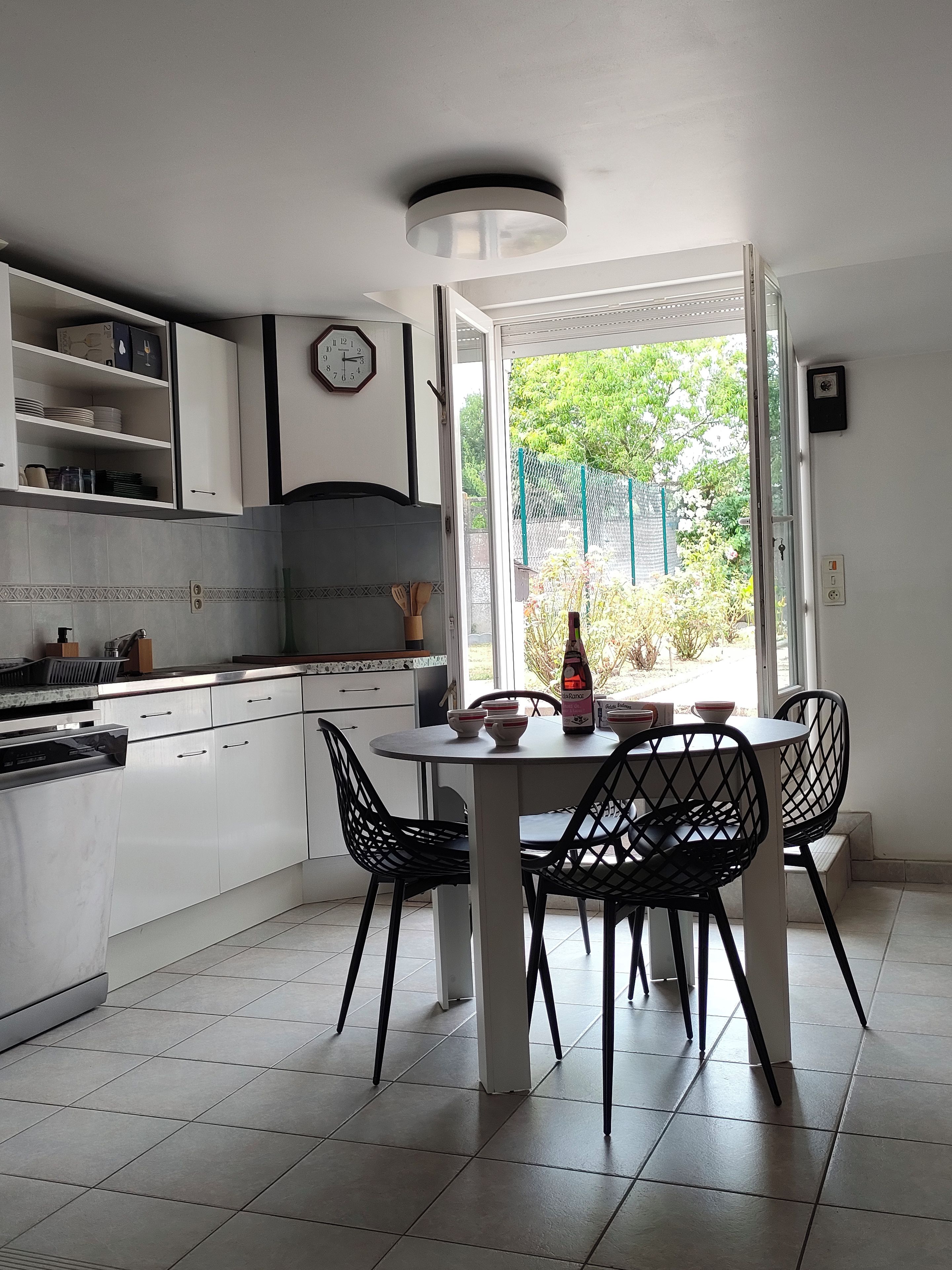 A kitchen with a dining table and chairs, a clock, and a view of greenery outside.