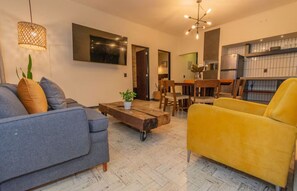 Sala de estar com sofá, mesa de centro e poltrona amarela. Há uma área de jantar com mesa e cadeiras, e uma cozinha com geladeira.