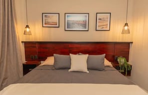 Cama bem arrumada com cabeceira de madeira, duas mesas de cabeceira e três quadros na parede.