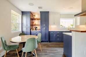 A modern kitchen with dark blue cabinets, a wooden dining table with green chairs, and a window with blinds. - Modern Oasis 3-bedroom house with AC in quiet Houston neighborhood (Houston)