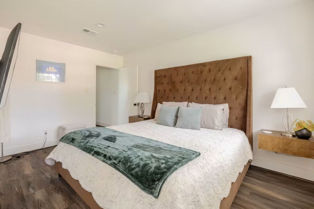 A bedroom with a large bed, a wooden headboard, two bedside tables with lamps, and a small wall-mounted TV.