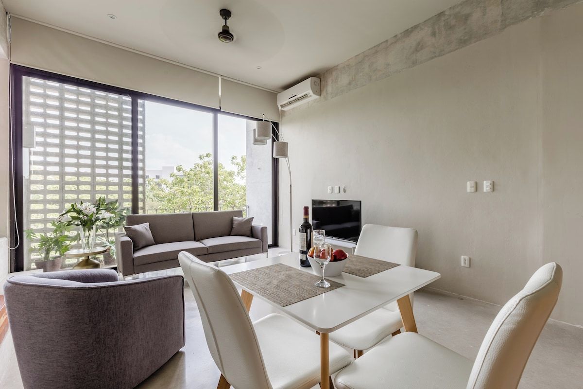A modern living room with a sofa, a dining table with chairs, a TV, and a large window with a view of greenery.