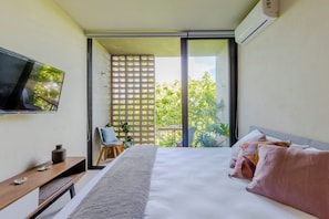 Un dormitorio con cama, televisor, escritorio y puerta corrediza de vidrio que da a un balcón con plantas.