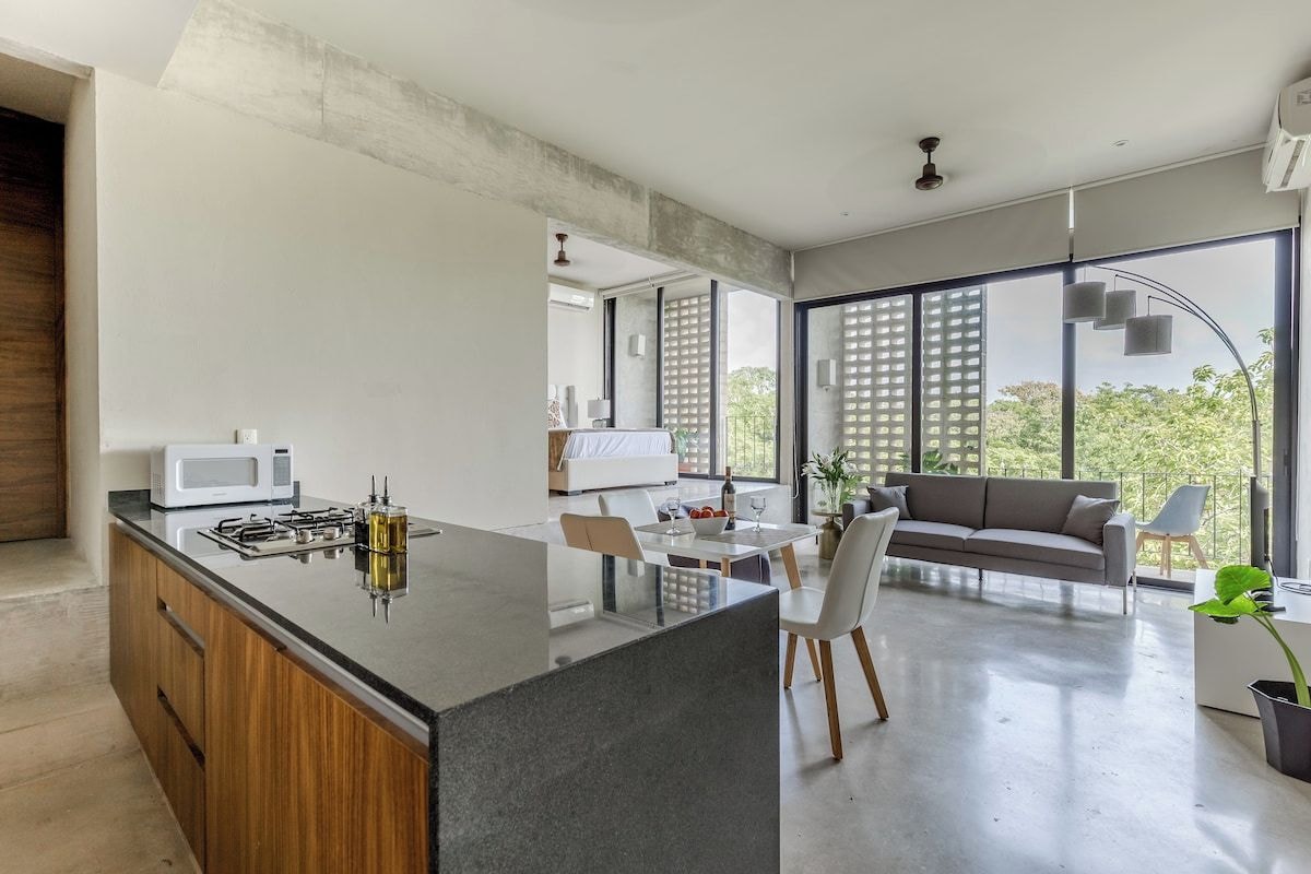 A modern kitchen with a central island, dining area with a sofa, and large windows offering a view of greenery.