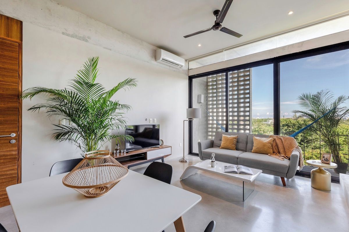 Ein modernes Wohnzimmer mit einer Couch, einem Couchtisch und einem Fernseher. Ein großes Fenster bietet einen Blick ins Freie.