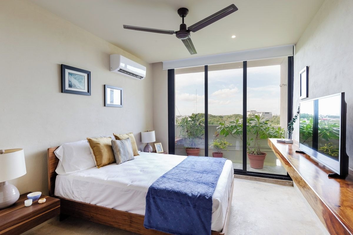 A bedroom with a bed, bedside tables, a ceiling fan, a large window, and a TV.