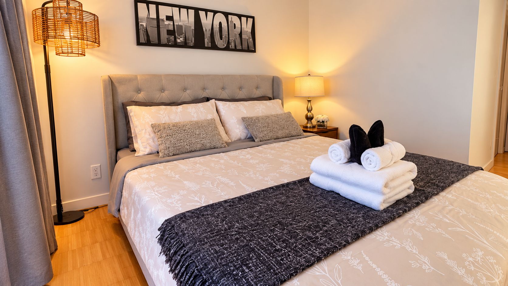 A hotel room with a bed, a wall-mounted lamp, a bedside table with a lamp, and a 'NEW YORK' sign.