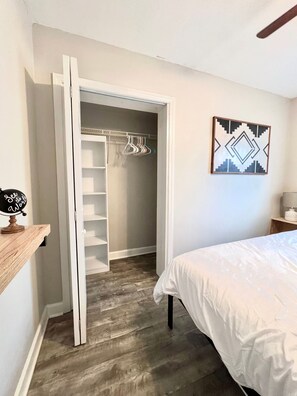 Quarto com cama, armário com roupas penduradas, prateleira de madeira e quadro na parede.