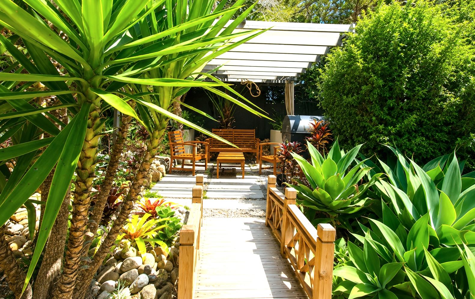 Una terraza de madera con zona de estar, rodeada de plantas tropicales y una pérgola.