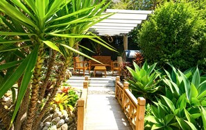 Una terraza de madera con zona de estar, rodeada de plantas tropicales y una pérgola.