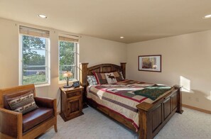 Ein Schlafzimmer mit einem großen Bett, einem Holznachttisch, einem Ledersessel und einem Fenster mit Blick auf Grünflächen.