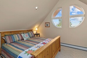 A bedroom with a sloped ceiling, a wooden bed, a nightstand with a lamp, and two windows with a view of the sky and trees.