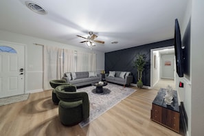 Uma sala de estar moderna com um sofá cinzento, duas poltronas verdes, uma mesa de centro e uma planta decorativa.
