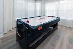 Uma mesa de bilhar com uma bola vermelha no canto, um armário preto e uma janela com cortinas brancas.
