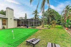 A backyard with a green artificial turf, a wooden table, a swing set, and a covered patio area. - Private Pool, Pickleball, Mini Golf, Game Room, Near AMI (Bradenton)