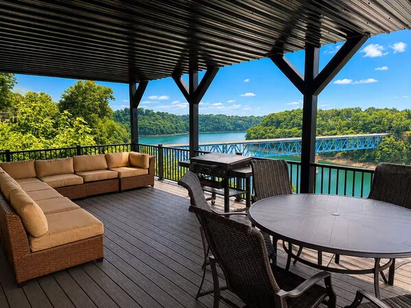 An outdoor deck with a sofa, chairs, and a table, overlooking a lake and a bridge.