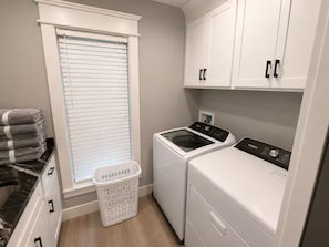 A laundry room with a front-loading washer and dryer, a window with blinds, a laundry basket, and a stack of folded towels. - Lake Cumberland Views by Lee’s Ford Marina 5BR, 3 Decks, Sleeps 10, Pet Friendly (Nancy)