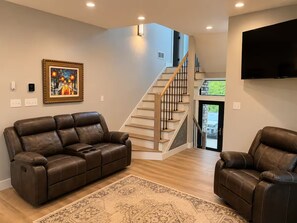 A modern living room with a brown leather sofa, a matching armchair, a patterned rug, a staircase, and a flat-screen TV mounted on the wall.