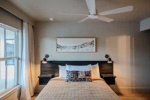 Un dormitorio con cama, ventilador de techo, mesita de noche, lámpara y ventana con cortinas.