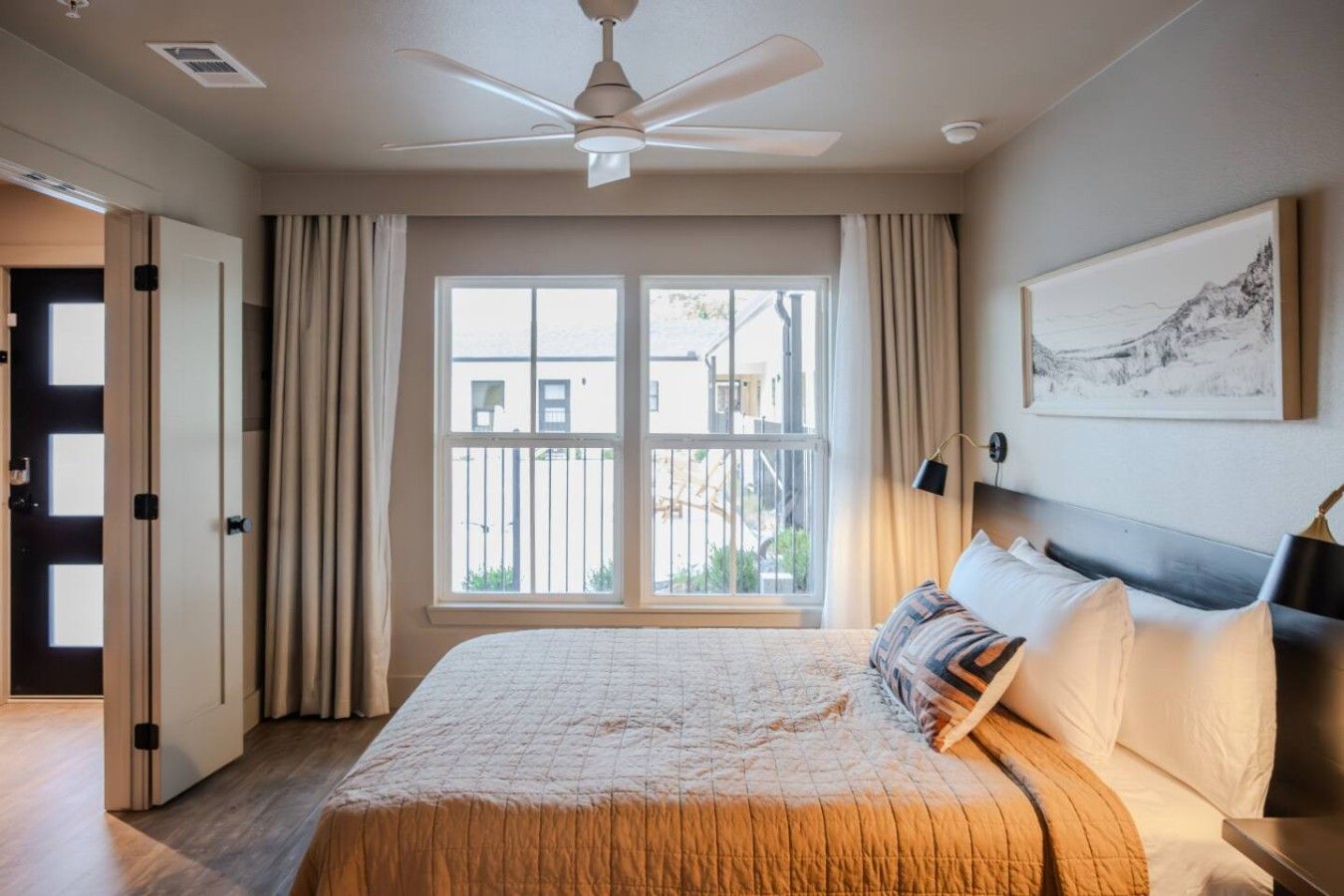 A hotel room with a bed, a ceiling fan, a large window with curtains, and a framed picture on the wall.
