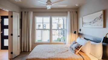 A hotel room with a bed, a ceiling fan, a large window with curtains, and a framed picture on the wall.