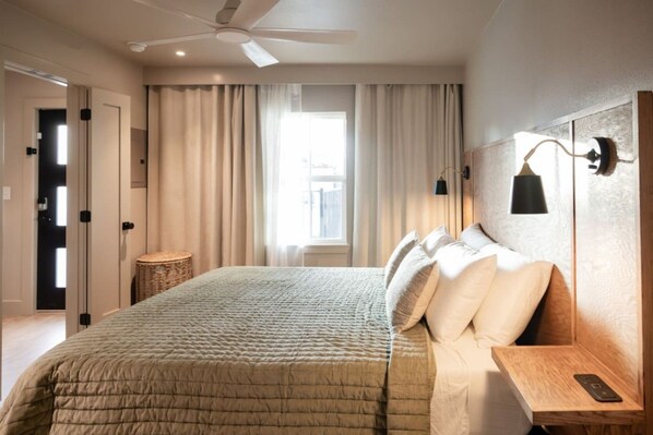 Ein Schlafzimmer mit einem Bett, einem Nachttisch, einer Lampe, einem Deckenventilator und einem Fenster mit Vorhängen.