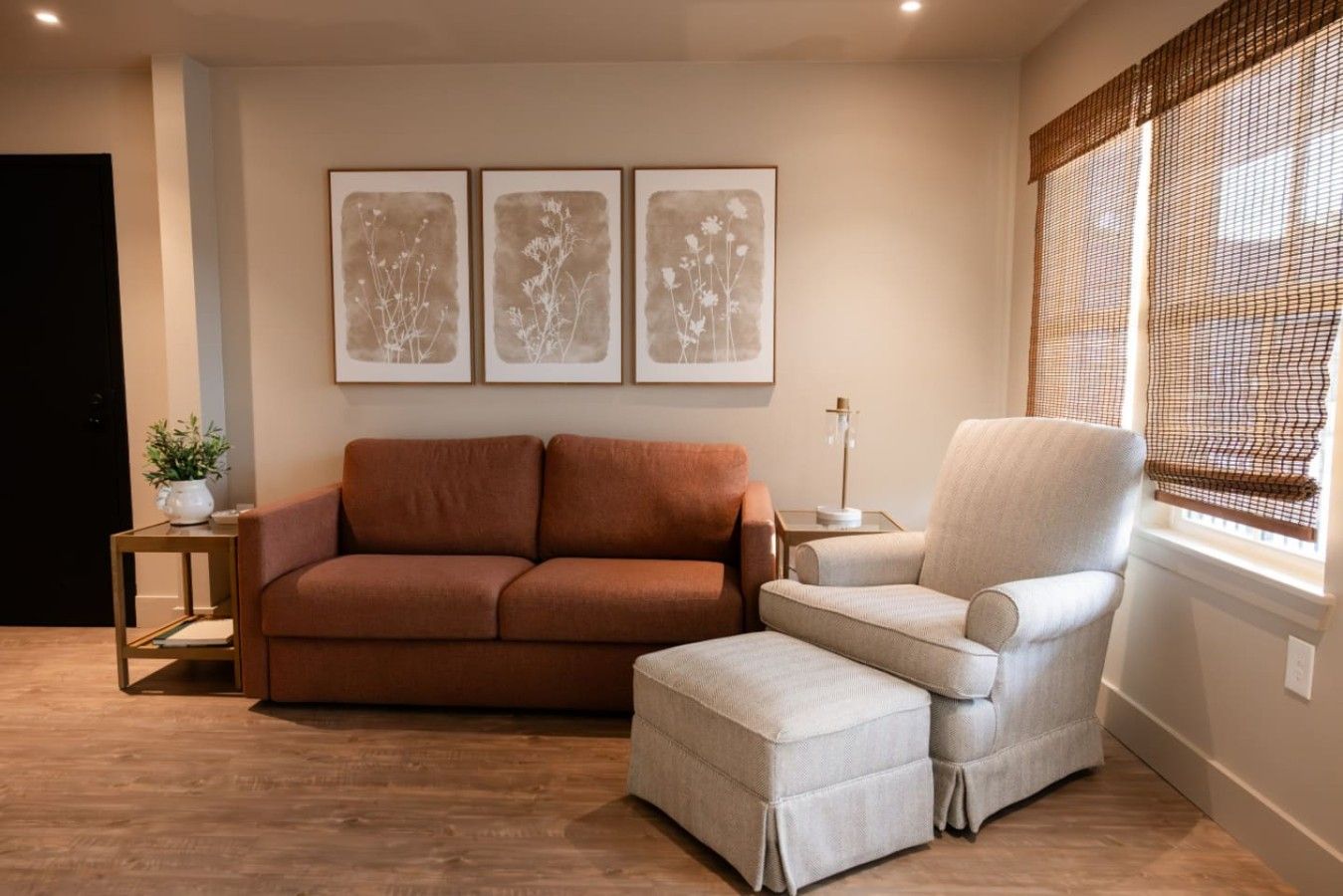 A living room with a brown sofa, a light-colored armchair, and a small table with a lamp. There are three framed pictures on the wall.