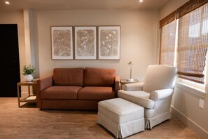 A living room with a brown sofa, a light-colored armchair, and a small table with a lamp. There are three framed pictures on the wall.