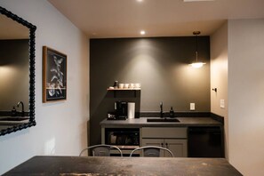 A compact kitchen with a dark countertop, a coffee machine, and a sink. There is a framed picture on the wall and a mirror with a beaded frame.
