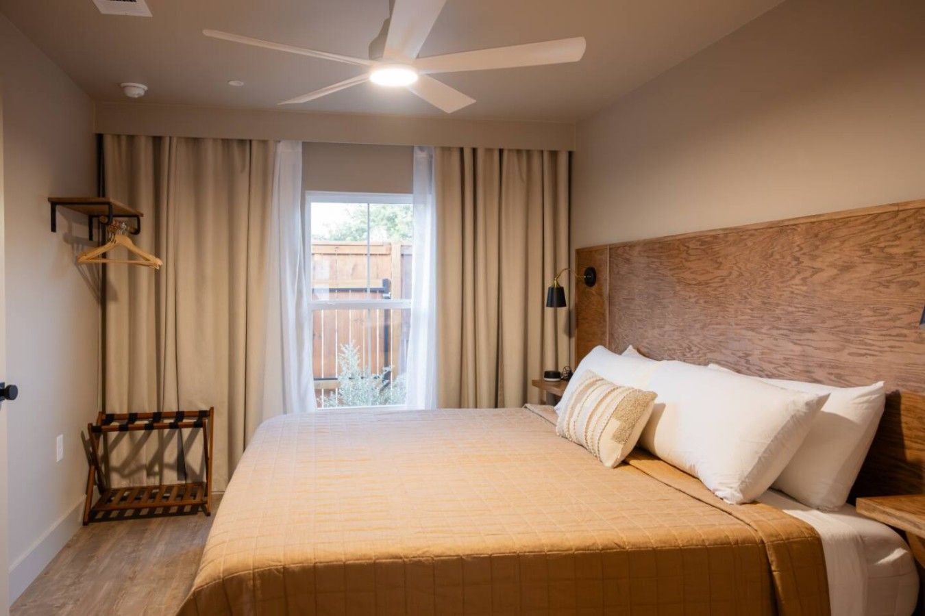 A hotel room with a bed, a ceiling fan, a window with curtains, a wooden headboard, and a nightstand with a lamp.