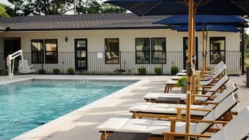 A poolside area with lounge chairs and umbrellas, a pool, and a building with windows and a fence.