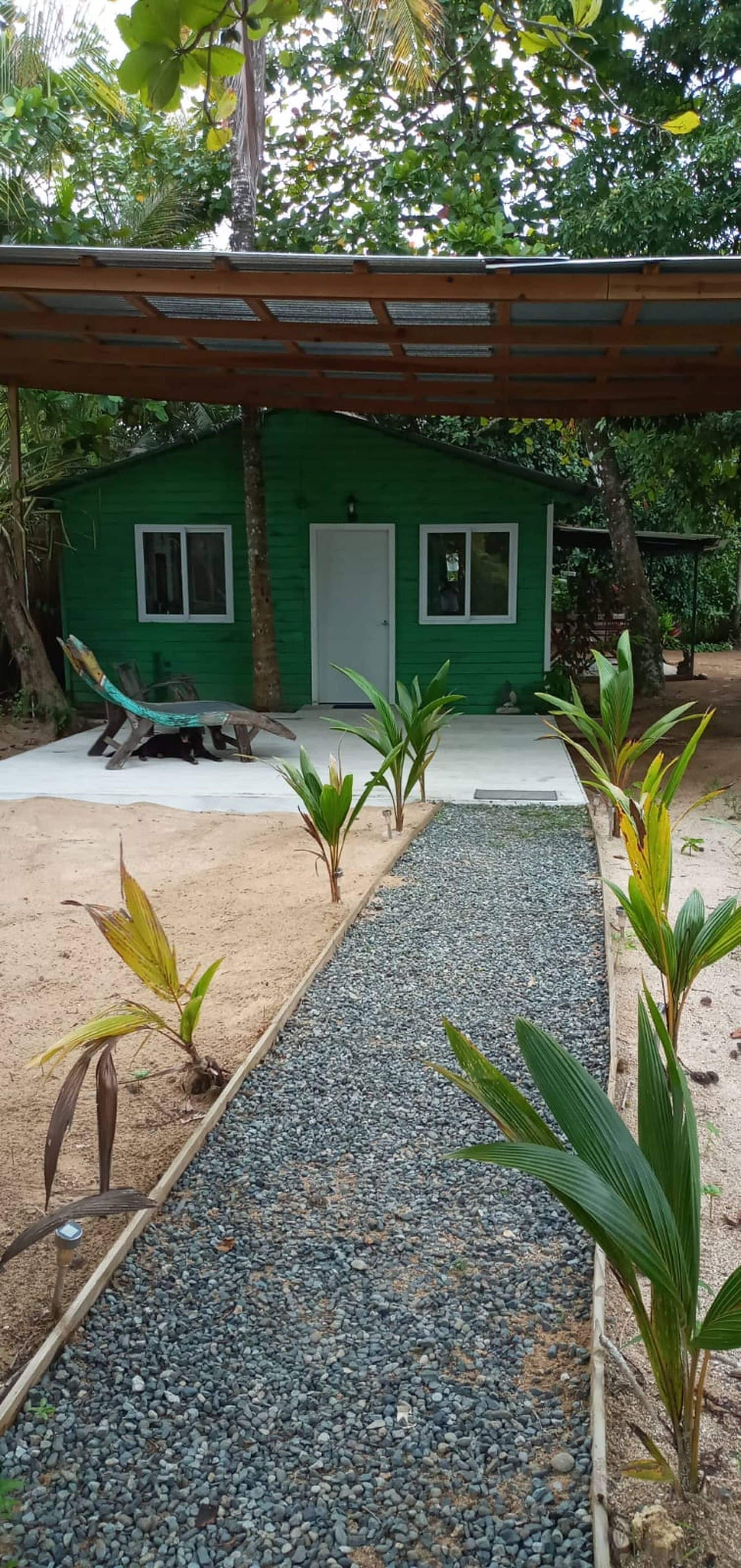 A green house with a gravel pathway leading to it, surrounded by tropical vegetation.